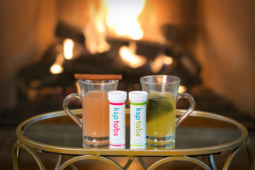 A tube of berry and lime KSPtabs sit next to two glasses of tea in front of a roaring fire.