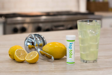 A tube of KSP lime next to some lemons and juicer. A glass of KSP lime is next to the tube.